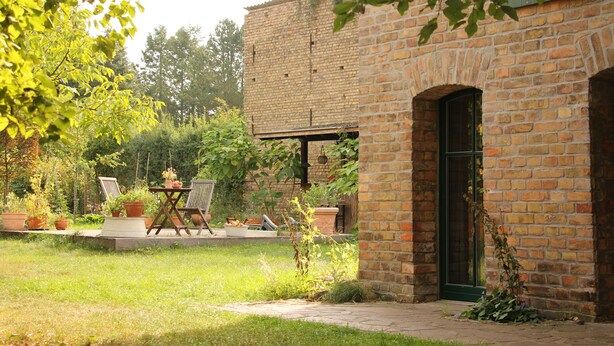 Edificio de ladrillo con puerta verde, un patio con mesa y sillas, y plantas en macetas.