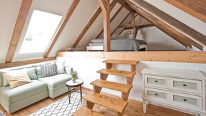 Un dormitorio tipo loft con techo inclinado, cama empotrada, sofá verde claro, una mesita redonda y un aparador blanco.