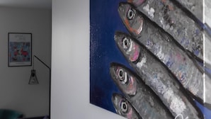 A room with a large painting of fish on the wall, a framed picture on the wall, a floor lamp, and a couch with a colorful pillow. - Apartment with patio, quiet, near the market and in the city center (Dinard)