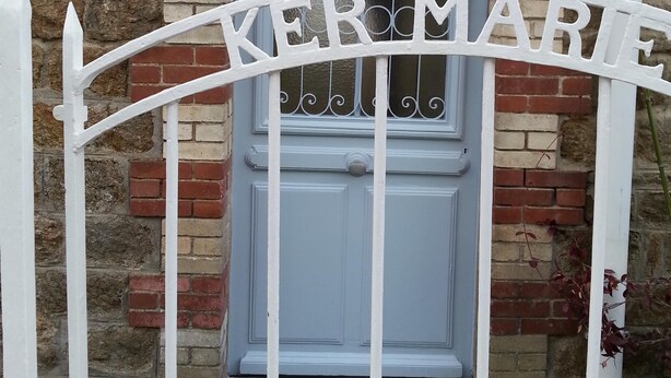 A white metal gate with the inscription "KERMAIRE" in front of a blue door with a glass panel, set against a brick wall. - Apartment with patio, quiet, near the market and in the city center (Dinard)