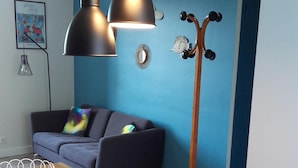 A modern living room with a blue accent wall, a wooden dining table, a black coat rack, and a gray sofa with colorful cushions. - Apartment with patio, quiet, near the market and in the city center (Dinard)
