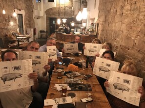 Un groupe de personnes est assis à une table dans un restaurant. Sur la table, on trouve des menus illustrés avec un dessin de cochon, ainsi que des verres à vin et des couverts.