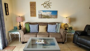 A living room with a brown sofa, a glass coffee table, and a painting on the wall. - Live the Beach Life! Stay in our FUN & AMAZING Townhome! (St Augustine)