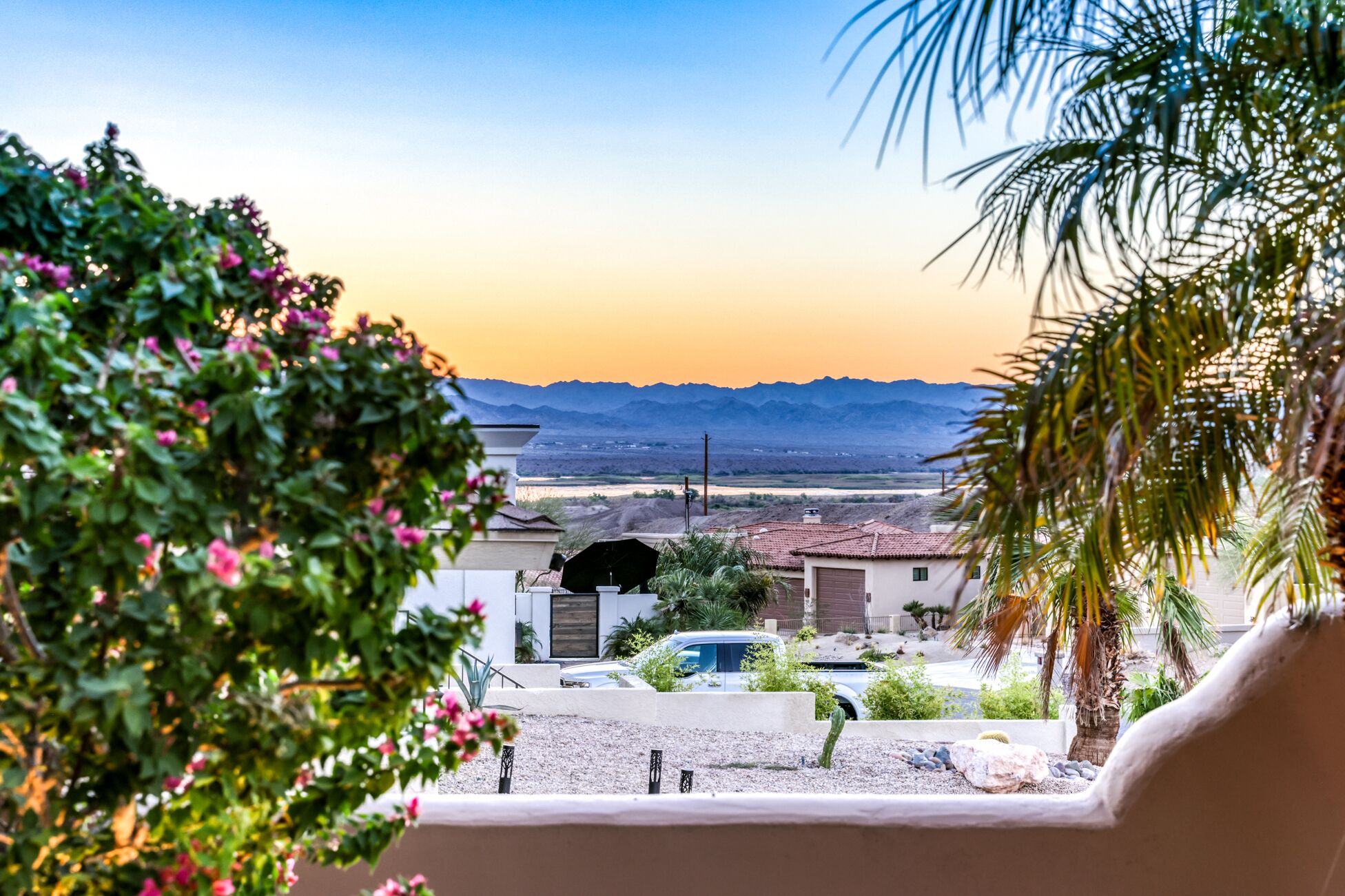 A residential area with a view of mountains in the distance, a white car parked in the driveway, and a cactus in the foreground.