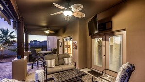 Un patio techado al aire libre con ventilador de techo, zona de estar con cojines estampados y un adorno en la pared.