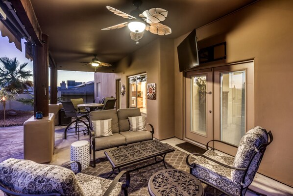 An outdoor patio area with a ceiling fan, seating arrangements, and a view of the outside.