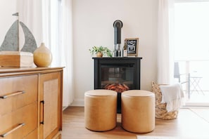 Une pièce chaleureuse avec un poêle à bois, une armoire en bois, deux poufs ronds, un panier en osier contenant une couverture et un panneau décoratif encadré.