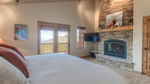 Une chambre avec un grand lit, une cheminée en pierre, un téléviseur fixé au-dessus, et une porte avec un cadre en bois.