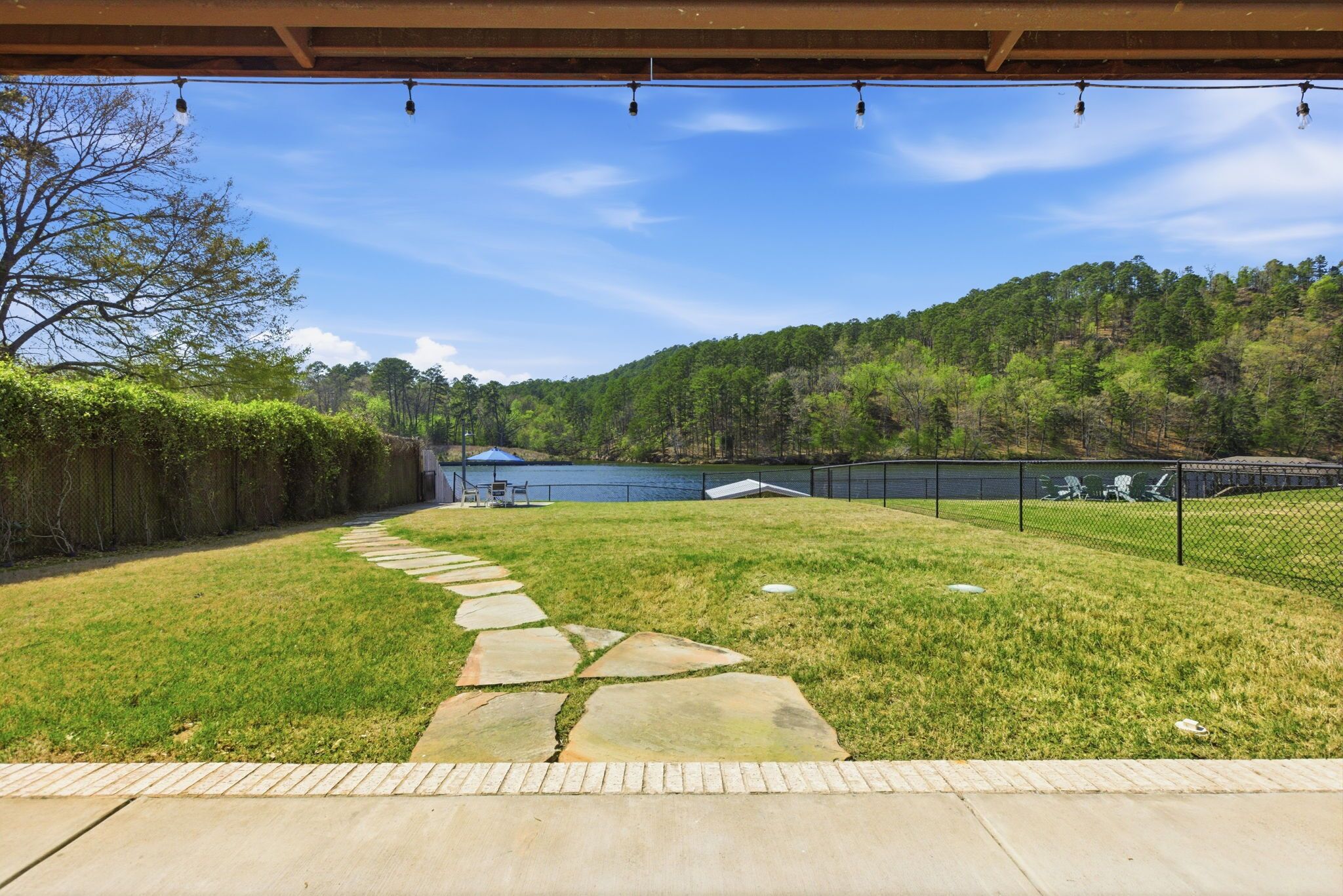 Un jardin avec un chemin de pierre, une zone délimitée par une clôture et une vue sur un lac et une colline boisée.