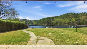 Un jardin avec un chemin de pierre, une zone délimitée par une clôture et une vue sur un lac et une colline boisée.