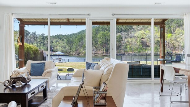 A spacious living area with a large window overlooking a lake and forest. - Stylish Lakefront Home - Last Minute Getaway for the Summer (Hot Springs)