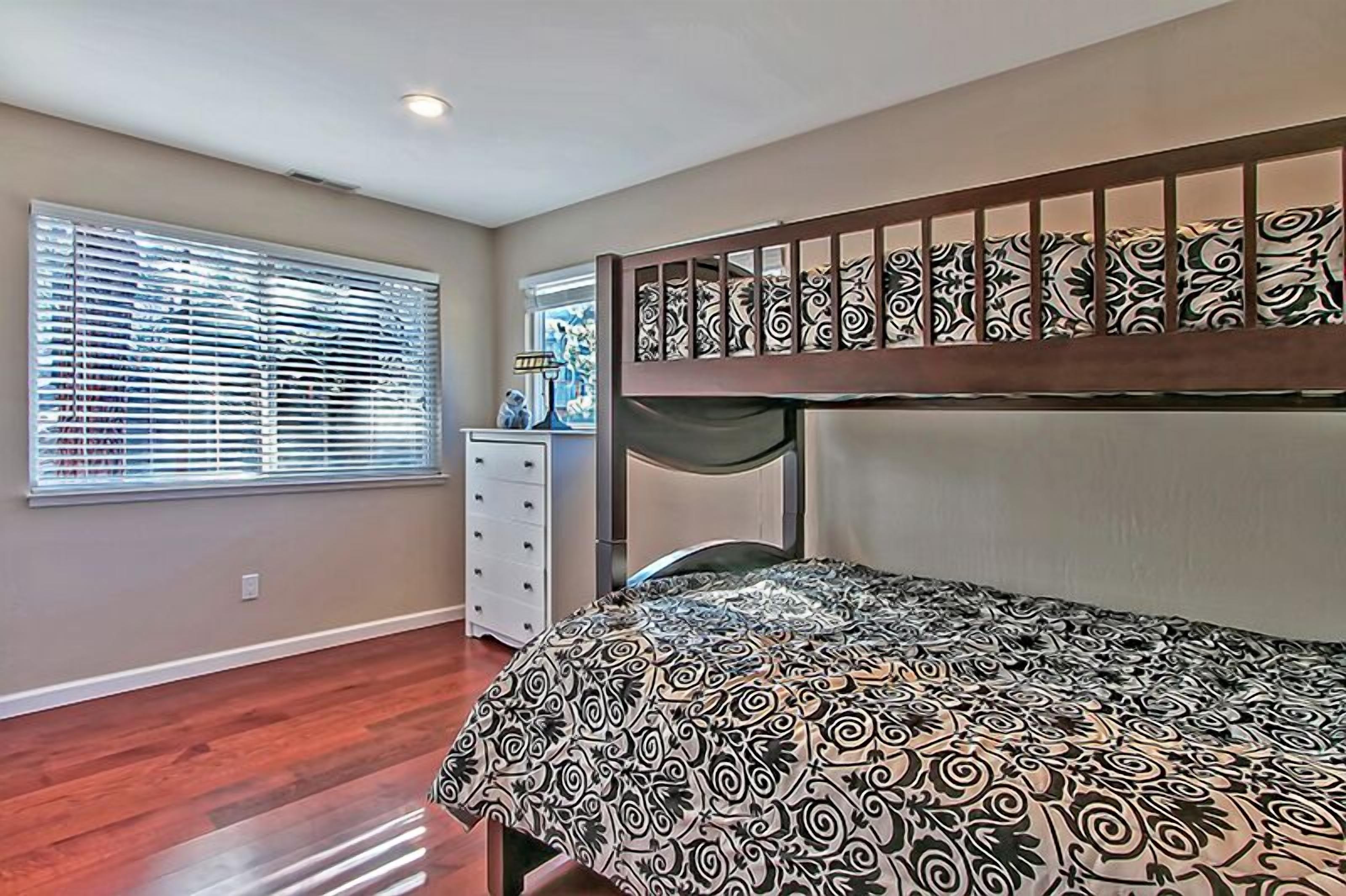 A bedroom with a bunk bed, a dresser, a window with blinds, and a patterned bedspread.