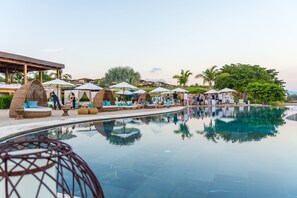 Un espace aménagé au bord de la piscine, avec des chaises longues et des parasols, une piscine à eau claire et un décor tropical agrémenté de palmiers.
