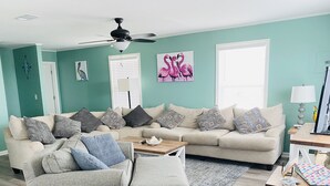 A living room with a sectional sofa, a loveseat, a coffee table, and a side table with a lamp. There is a ceiling fan, a wall clock, and a window with blinds.