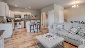 A modern kitchen with white cabinets, a stainless steel refrigerator, and a microwave. There is a dining area with a grey sofa and a small table. - Razorback Getaway Close to University!  (Fayetteville)