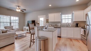 A modern kitchen with white cabinets, a granite countertop, and a built-in island. There is a dining area with a sofa and chairs, and a ceiling fan above. - Razorback Getaway Close to University!  (Fayetteville)