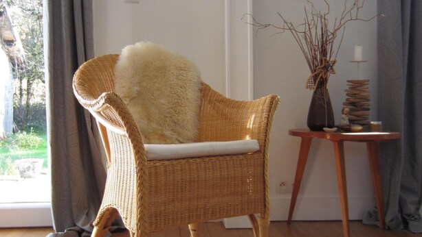 Una silla de mimbre con un cojín mullido en una habitación con piso de madera, una decoración con forma de corazón en la pared y una pequeña mesa con un florero y fotografías.