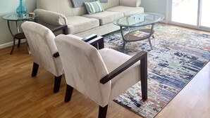 A living room with a sofa, two armchairs, a glass coffee table, a ceiling fan, and a large window with blinds. - ☀ Breezy, Open Condo, Steps from Beach and Boardwalk with Ocean Views & Pool ☀  (Carolina Beach)