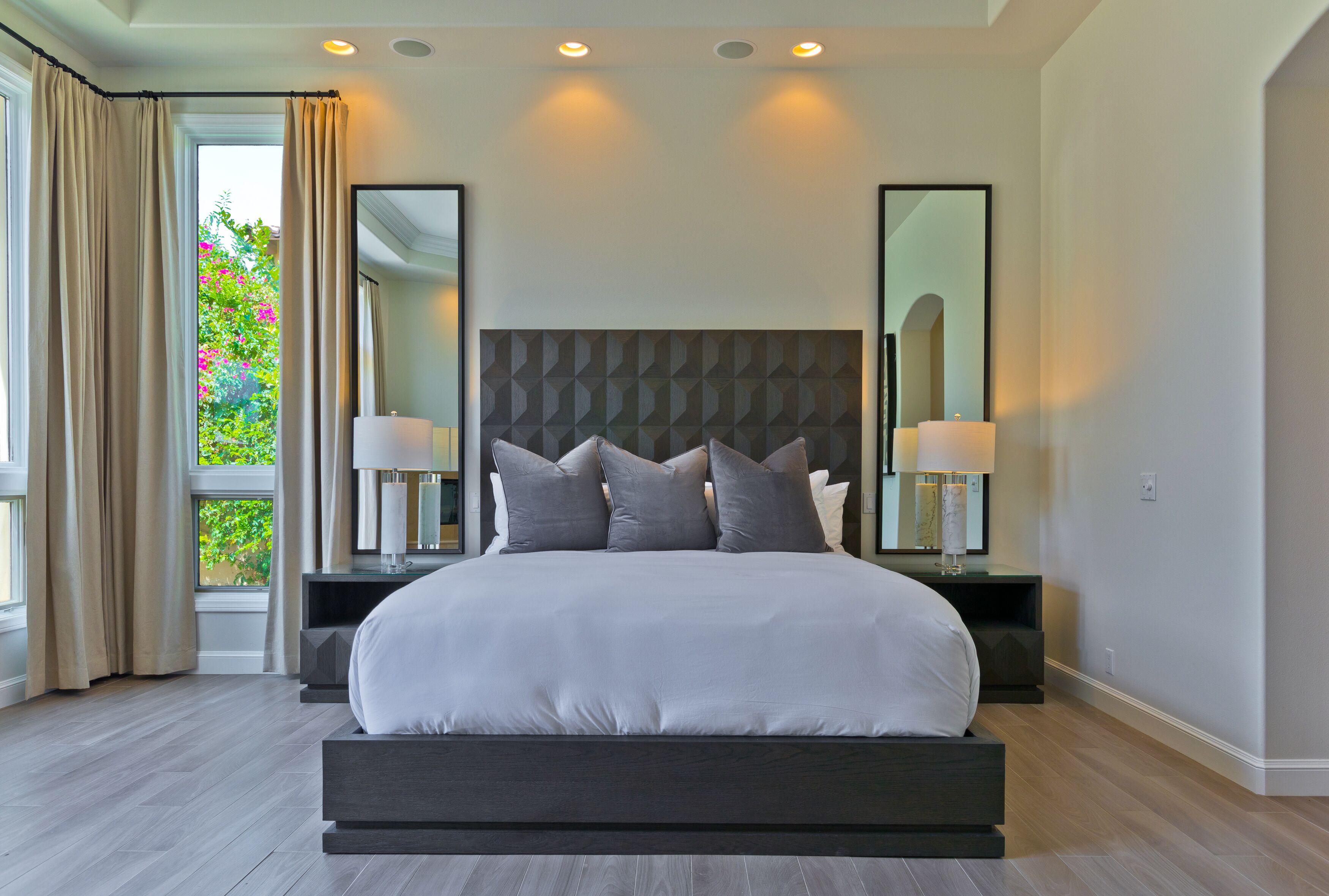 A modern bedroom with a large bed, two bedside tables with lamps, a geometric-patterned headboard, and large mirrors on the walls.