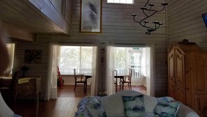 Una habitación espaciosa con paredes de madera, un ventilador de techo, una zona de comedor con sillas y una mesa, y una cama con ropa de cama estampada.