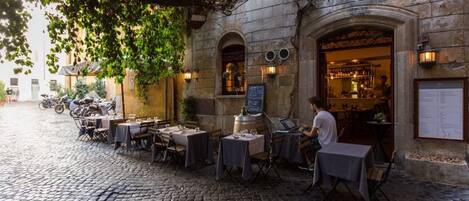 An outdoor café with tables and chairs, a stone building with an arched entrance, and a cobblestone street.