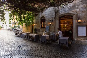 An outdoor café with tables and chairs, a stone building with an arched entrance, and a cobblestone street.