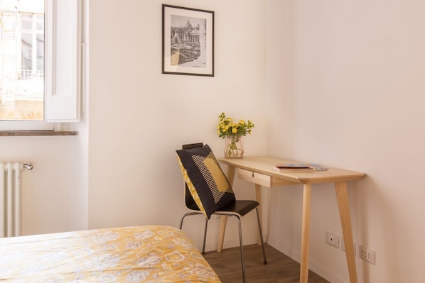 A bedroom with a bed, a desk, a chair, a radiator, a window, and a framed picture on the wall.