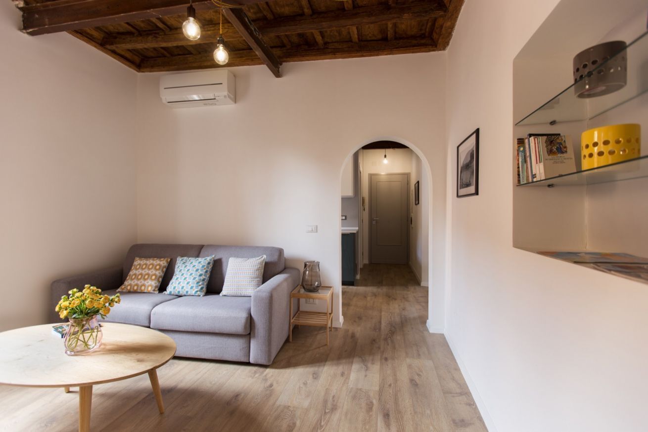 A modern living room with a grey sofa, a round wooden coffee table, and a vase of flowers. There is a bookshelf with books and decorative items, and an arched doorway leading to another room.