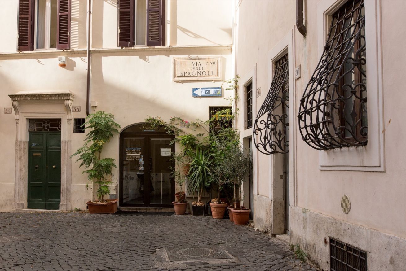 Eine enge Kopfsteinpflasterstraße mit Hauseingang, Blumentöpfen und einem Schild mit der Aufschrift 'VIA R. VIII DEGLI SPAGNOLI'.