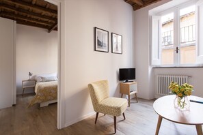 A room with a bed, a chair, a TV, and a window with a view of buildings.