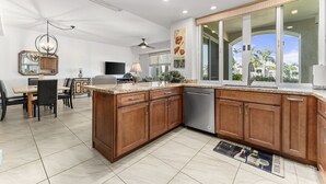 A modern kitchen with wooden cabinets, a dishwasher, and a dining area with a table and chairs. - #1301 Ocean Views, 1st Floor, Best location, Courtyard, Great Promotion (Kihei)