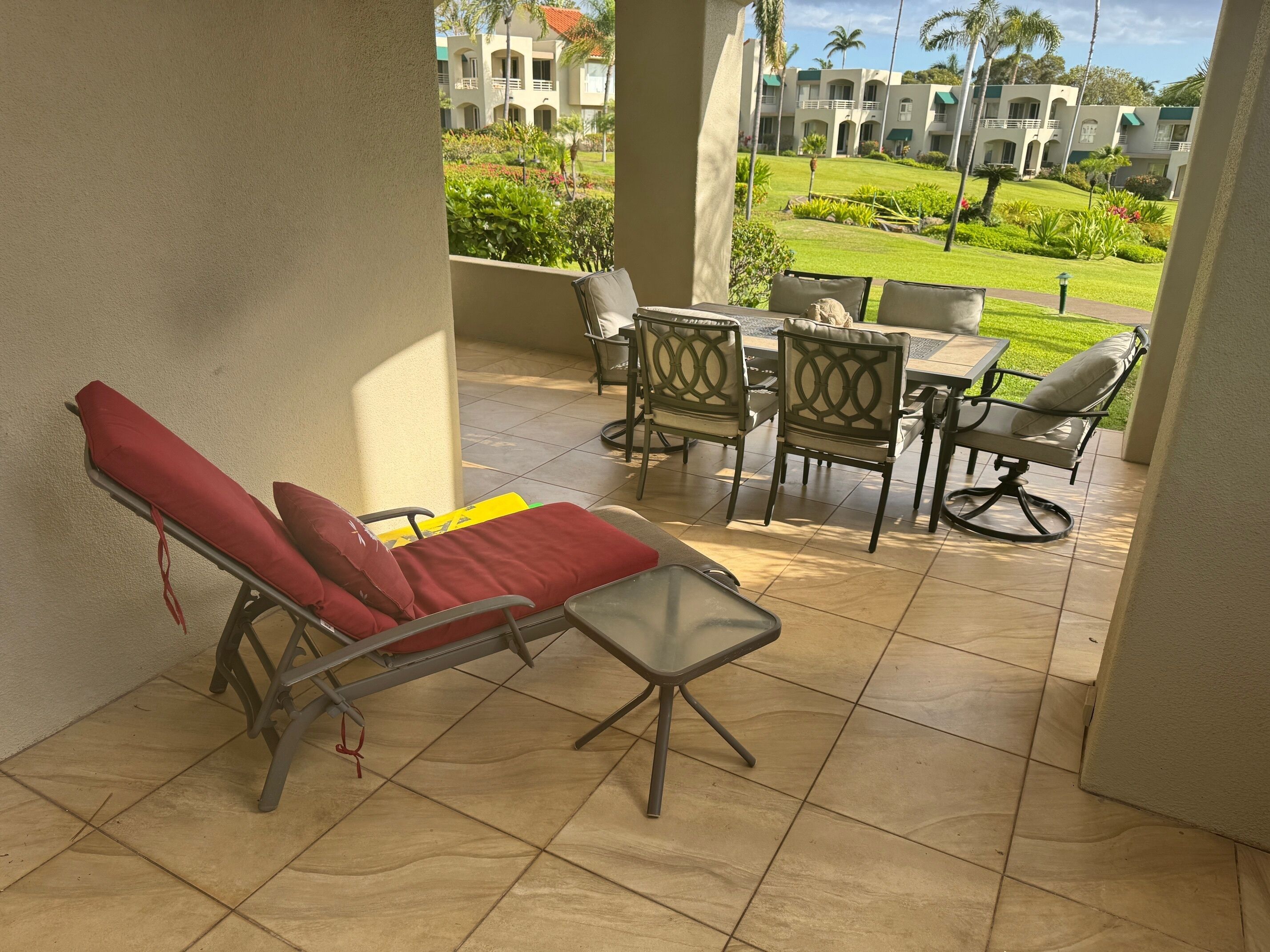 A covered outdoor seating area with lounge chairs and a table, overlooking a well-maintained garden and a building with multiple balconies.