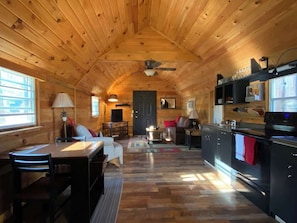 A cozy cabin interior with a wooden ceiling, a dining area, a kitchen with a stove, and a seating area with a fireplace. - Cozy Cabin for 2 in the Heart of RRG! (Campton)