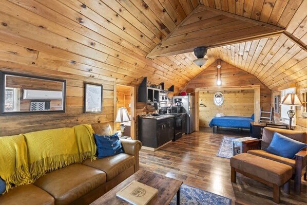 A cozy log cabin interior with a wooden ceiling, a living room with a sofa and armchair, a kitchen area, and a bedroom with a bed. - Cozy Cabin for 2 in the Heart of RRG! (Campton)