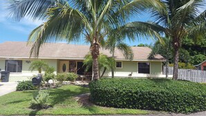 A single-story house with a beige exterior, a brick roof, and a well-maintained front yard with palm trees and a hedge.
