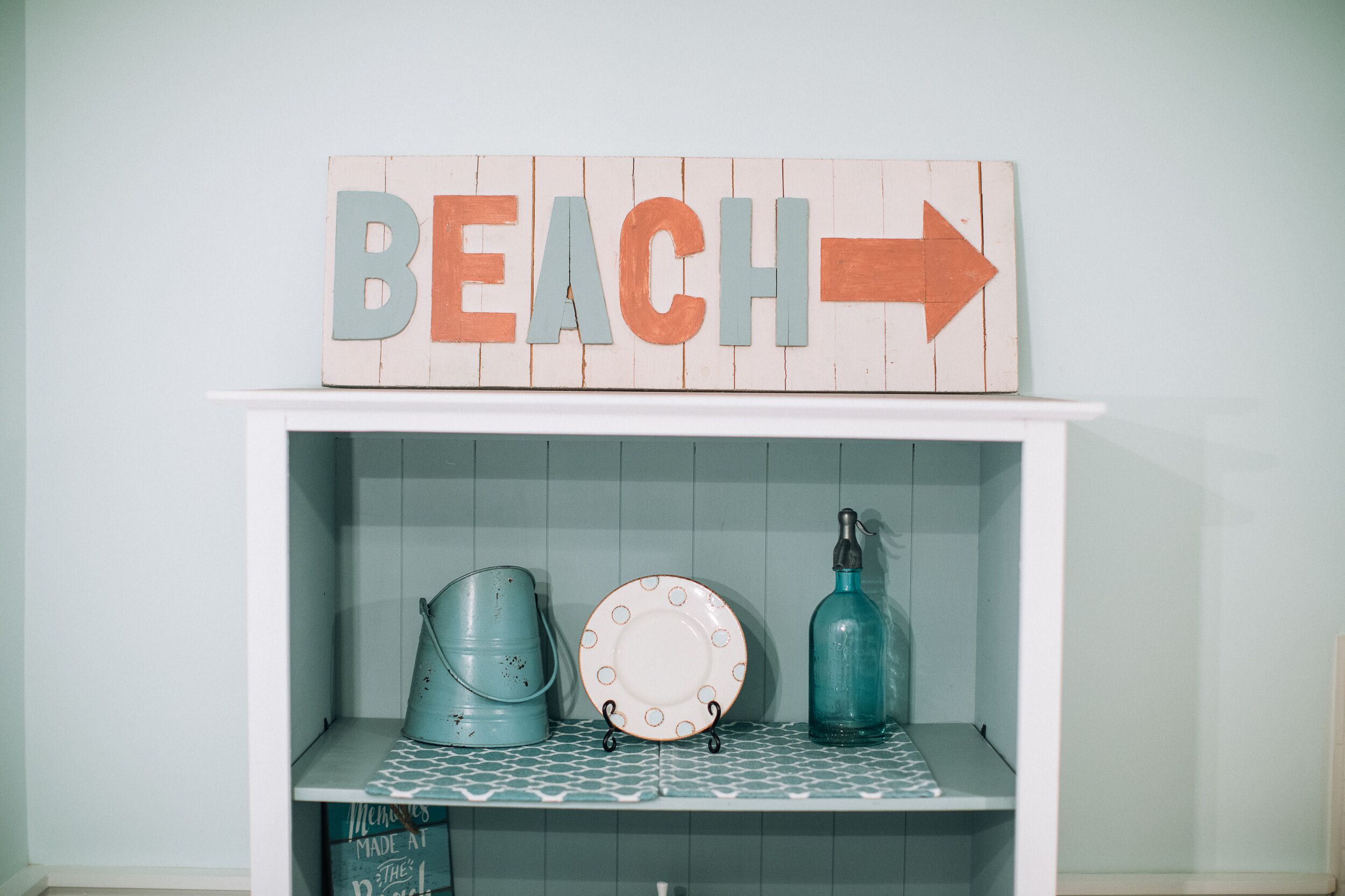 A shelf with a 'BEACH' sign, a blue bucket, a white plate, and a blue bottle.
