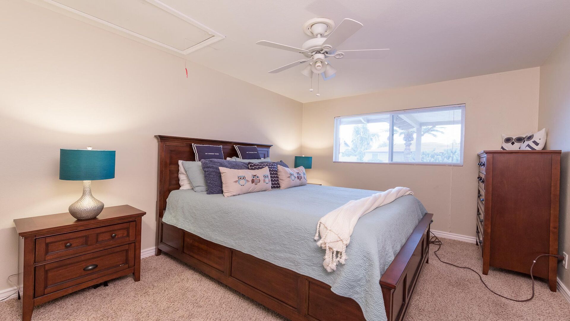 A bedroom with a bed, two bedside tables, a nightstand, a ceiling fan, a window with blinds, and a wardrobe.