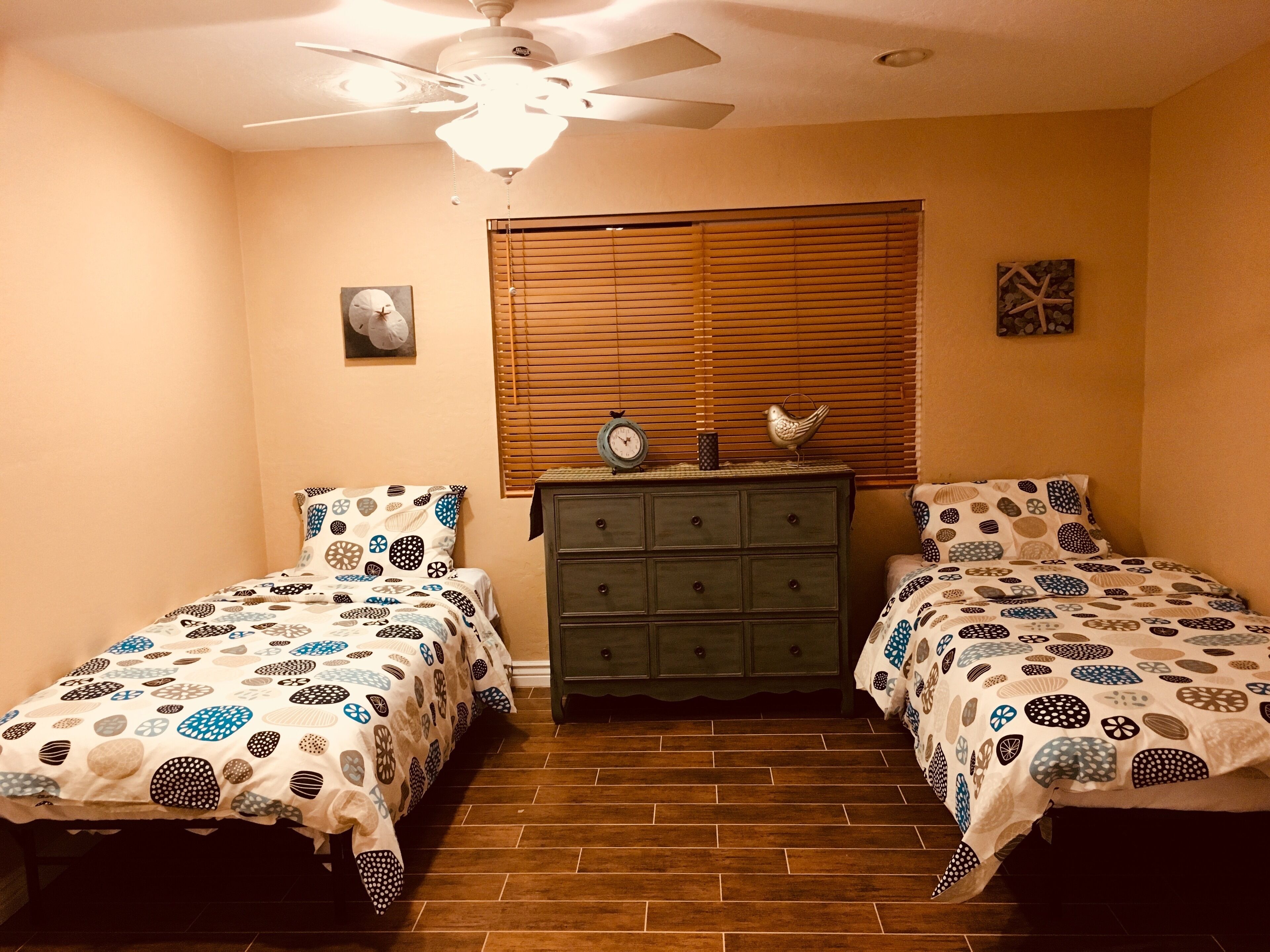 Une chambre avec deux lits, une commode, un ventilateur de plafond et une fenêtre avec des stores.
