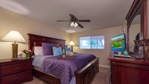 Une chambre à coucher comprenant un lit, deux tables de chevet, une commode, un ventilateur de plafond, une fenêtre et un téléviseur.