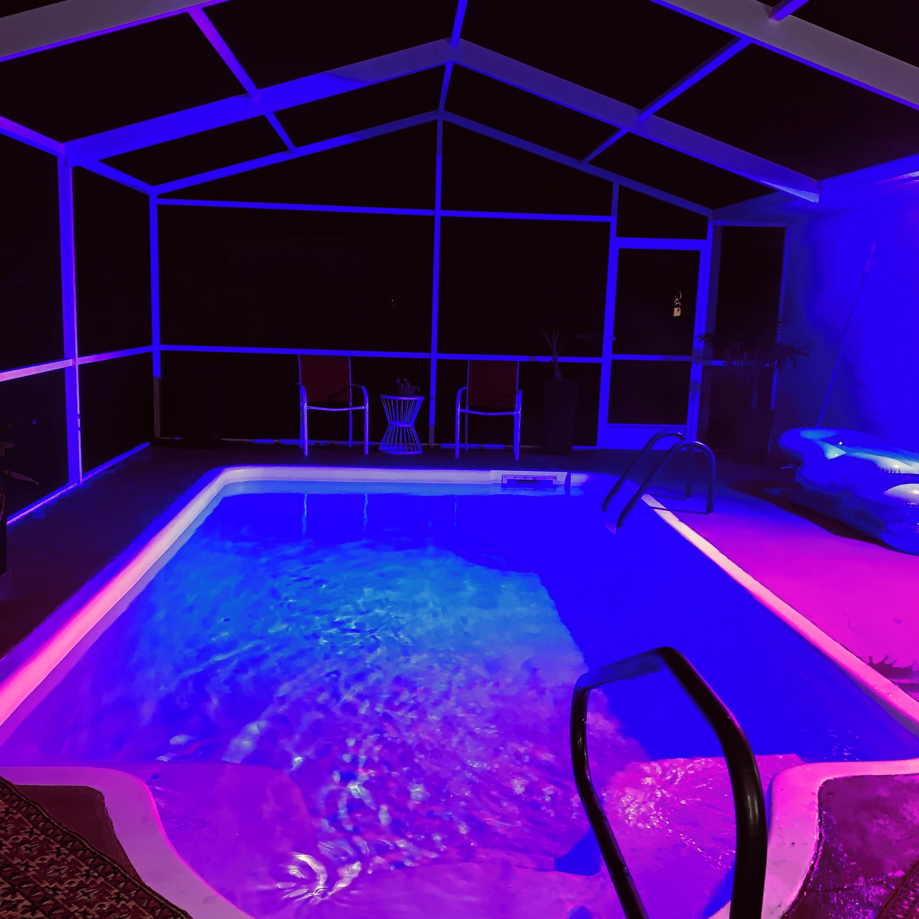 An indoor swimming pool with a geometric ceiling structure, surrounded by chairs and a small table.