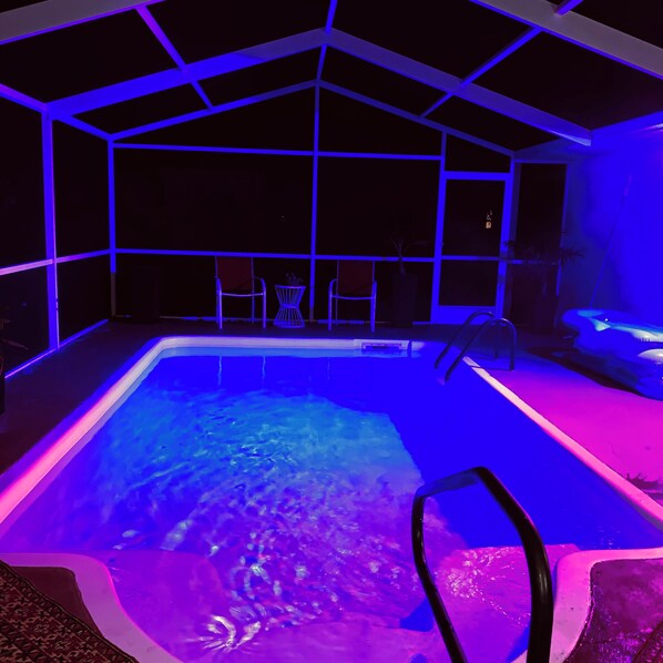 An indoor swimming pool with a geometric ceiling structure, surrounded by chairs and a small table.
