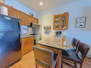A compact kitchen with a refrigerator, stove, microwave, and wooden cabinets. There is a dining table with chairs and a wine bottle.