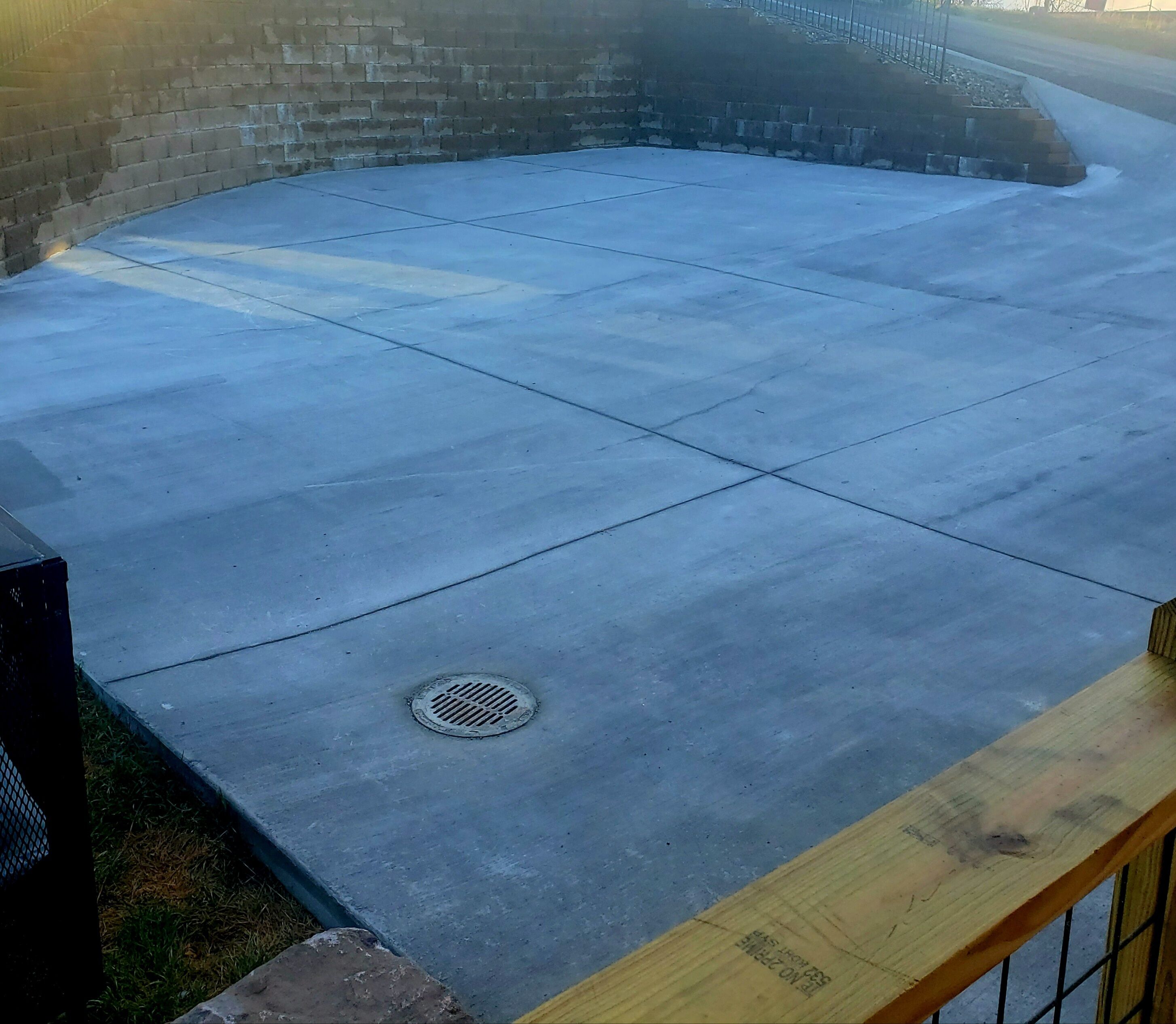 Une terrasse en béton avec un drain en forme de grille métallique, entourée d’un mur de briques et d’une rambarde en bois.