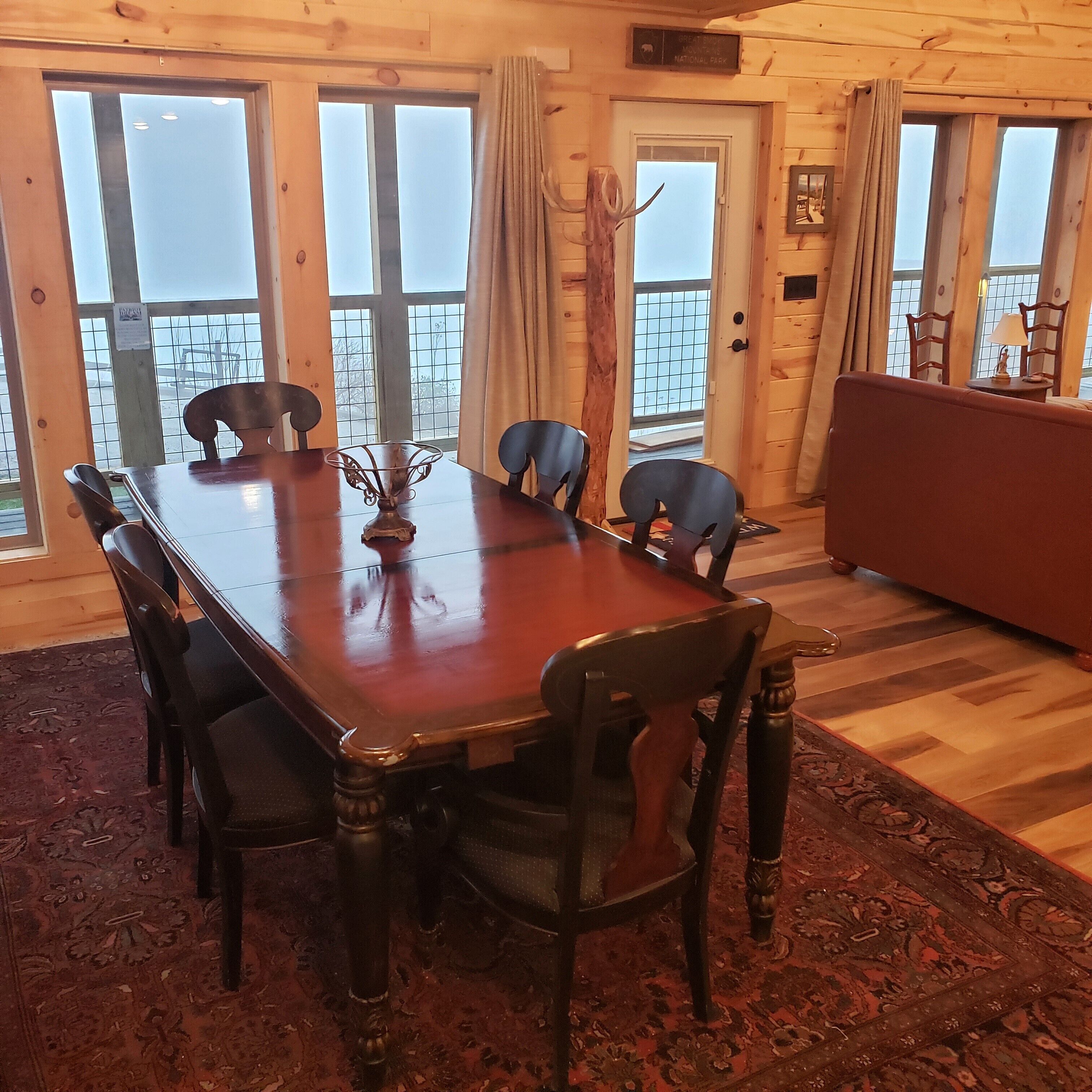 Une salle à manger en bois, avec une grande table et des chaises, un vase décoratif, et une vue sur un paysage enneigé par de grandes fenêtres.