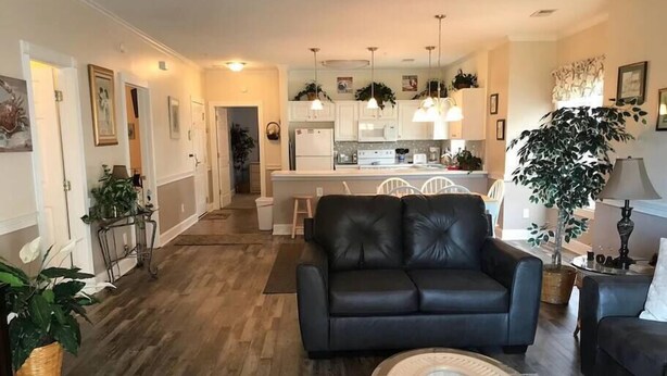 A modern living room with a black leather sofa, a glass coffee table, and a dining area with white cabinets and pendant lighting. - Charming 3 Bedroom Condo in Myrtlewood (Myrtle Beach)