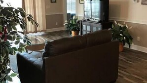 A living room with a dark-colored sofa, a television on a wooden cabinet, and a ceiling fan. There are two potted plants and a glass coffee table. - Charming 3 Bedroom Condo in Myrtlewood (Myrtle Beach)
