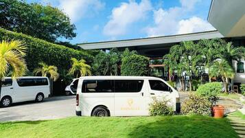 A white van parked in front of a building with a covered entrance, surrounded by lush greenery and palm trees.