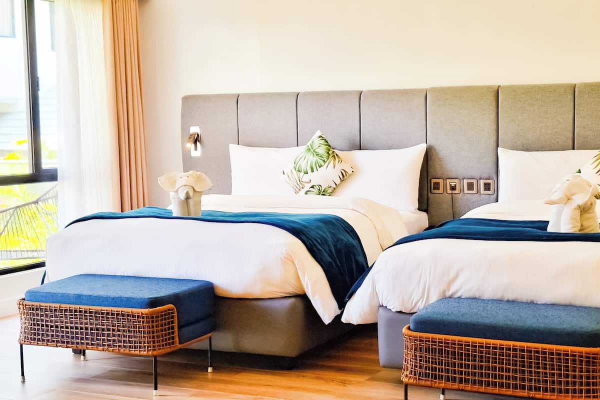 Un letto matrimoniale con testiera grigia, biancheria da letto bianca e cuscini decorativi. Due pouf in vimini e un tavolino con un vaso bianco.