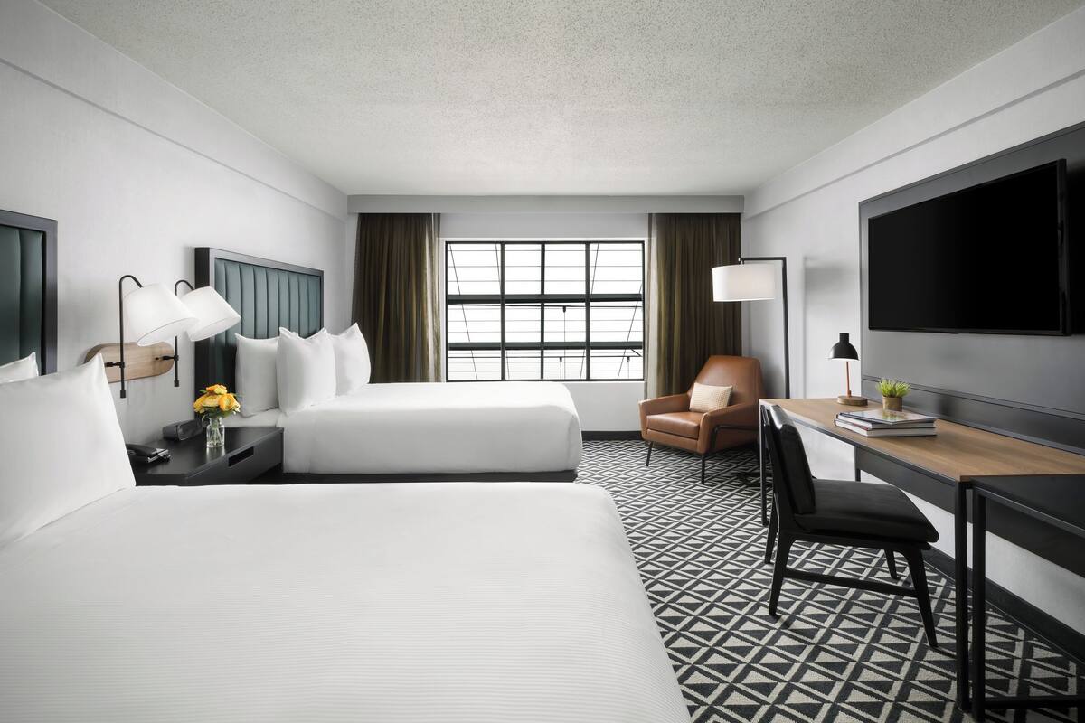 A hotel room with two beds, a desk, a chair, a TV, and a window with curtains.