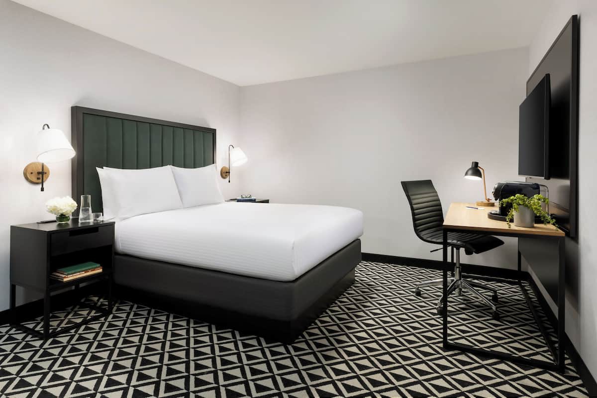 A modern hotel room with a large bed, a desk with a chair, a flat-screen TV, and a patterned floor.